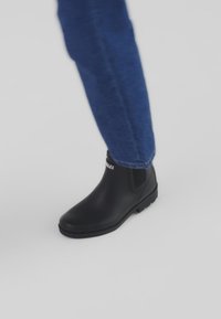 Czarny gumowy botek za kostkę z elastycznymi panelami bocznymi i teksturowaną podeszwą, zestawiony z niebieskimi dżinsami. Skupienie na funkcjonalności i designie.