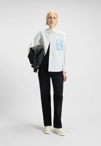 Witte button-up shirt met blauwe grafiek op de linkerborst, gecombineerd met zwarte broek en een zwarte rugzak, met crègekleurige sneakers.
