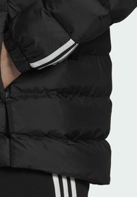 Veste matelassée noire avec un motif matelassé, comprenant des accents rayés blancs sur les poignets et une poche zippée sur le côté.