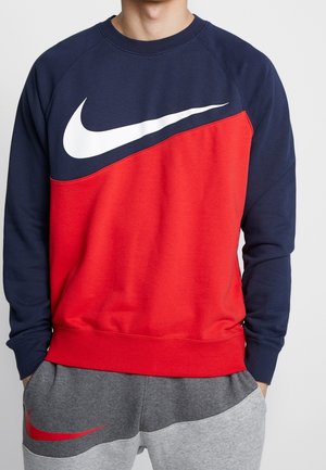 Mężczyzna ubrany w granatową i czerwoną bluzę z dużym białym logo Nike oraz szare spodnie dresowe z czerwonym logo Nike.