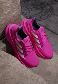 Lyserøde Adidas løbesko med hvide striber, der hviler på stenetrin nær en metaldæksel på fortovet.
