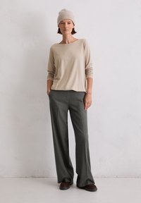 Beige langærmet top med vid halsudskæring, parret med løstsiddende grå bukser og brune sko. En strikket beanie fuldender outfittet.
