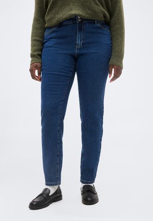 Persona con jeans blu a vita alta, maglione verde oliva, calzini bianchi e mocassini in pelle nera in piedi su sfondo bianco.