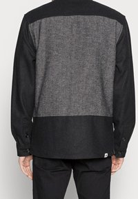 Sort flannel skjorte med et tekstureret gråt bagpanel. Har lange ærmer og knaplukninger ved manchetterne, samt en solid sort bundkant.