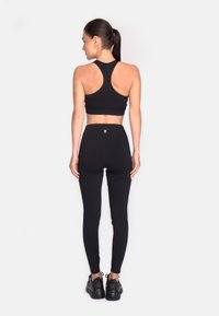 Ženska z temnim konjškim repom, oblečena v črno športno modrček, hlače z visokim pasom in masivne črne teniske, stoji s hrbtom obrnjena proti beli ozadju.