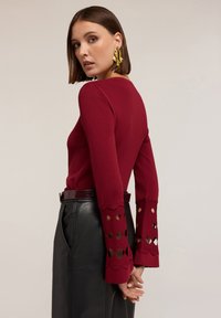 Femme aux cheveux bruns courts portant un haut rouge à manches longues avec des découpes aux manches et une jupe en cuir noire, tournant le dos avec les mains jointes.