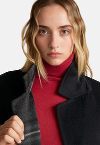 Abrigo de lana negro con solapa de muesca, que presenta una textura suave. Debajo, se puede ver una blusa de cuello alto roja ajustada. Vista cercana del área del cuello.
