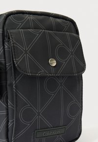 Must Calvin Kleini kott geomeetrilise joonemustriga, esiküljel tasku korgiga, mis on kinnitatud metallist klõpsnupuga, ja alumisel esiküljel logoplaat.