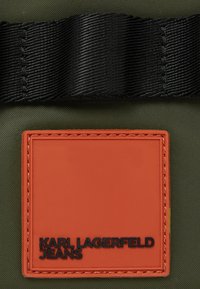 Olivegrüner Stoff mit einem schwarzen strukturierten Riemen und einem orangefarbenen Gummiknopf mit eingeprägtem schwarzen Text "KARL LAGERFELD JEANS."