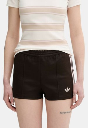 Donna che indossa pantaloncini neri Adidas con strisce bianche e logo, abbinati a una maglietta a maniche corte a righe bianche e beige.