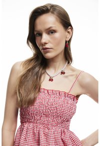 Rood-wit geruit jurkje met een smock-borstvoeding en dunne bandjes. Sieraden bestaan uit een parelketting en rode kers oorbellen.