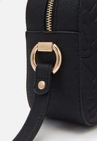 Sort lædertaske med præget logo, guldfarvet hardware, lynlås lukning og aftagelig skulderrem. Struktureret overflade og rund ring som accent.