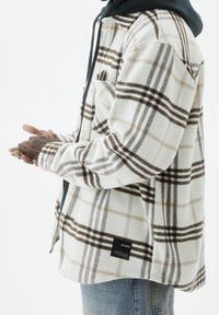 Camicia in flanella a quadri bianca e beige con strisce marroni, dotata di taschino sul petto e etichetta nera, indossata sopra un felpa scura.