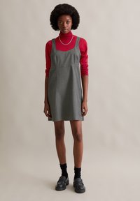 Graues Nadelstreifen-Kleid mit quadratischem Ausschnitt und A-Linie, über einen roten Rollkragenpullover gelegt; kombiniert mit schwarzen klobigen Loafers und Socken.