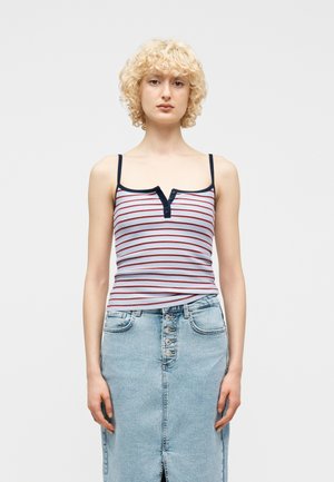 Άτομο με ξανθά σγουρά μαλλιά, που φοράει αμάνικο ριγέ τοπ και τζιν φούστα ψηλόμεση με κουμπιά μπροστά, στέκεται μπροστά σε λευκό φόντο.