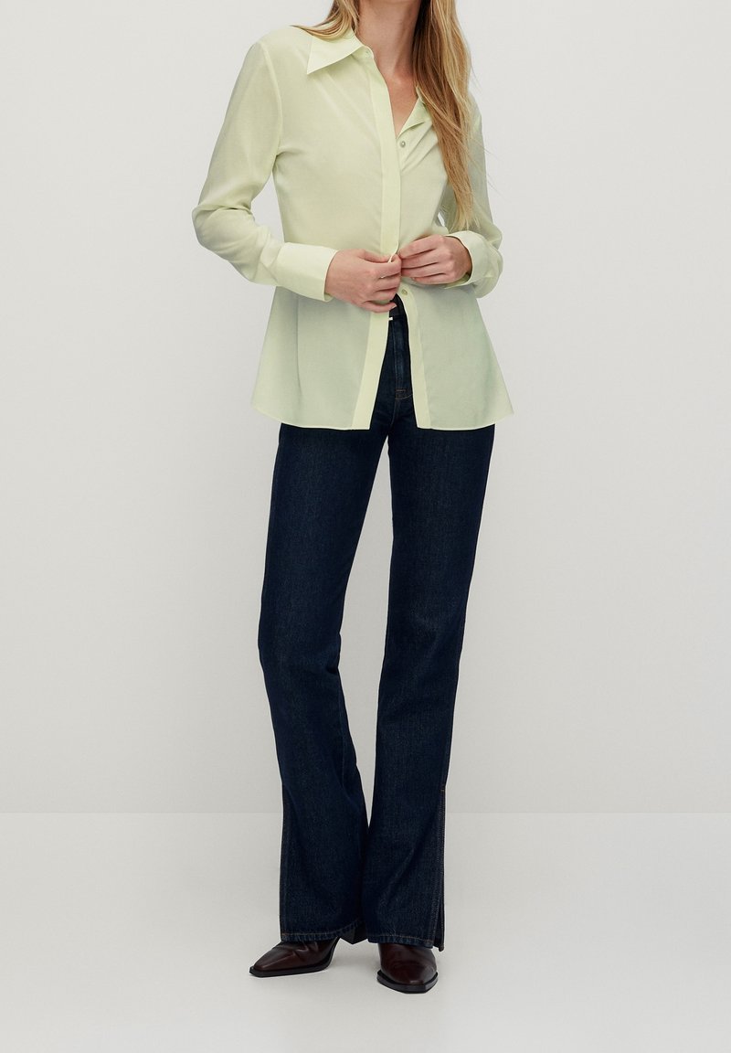 Camicia button-up gialla chiara con colletto a punta e maniche lunghe, abbinata a jeans svasati blu scuro e scarpe marroni.