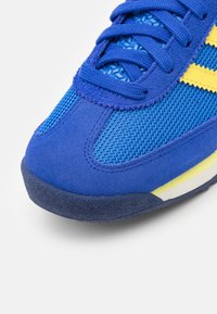 Blue athletic shoe with mesh and suede materials, featuring yellow accents, textured tongue, and rubber sole. Close-up of toe area.