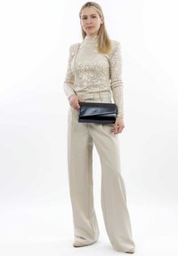 Top beig de lentejuelas con mangas largas y cuello alto; clutch negro con diseño plegable; pantalones crema de pierna ancha; zapatos beigs de punta.