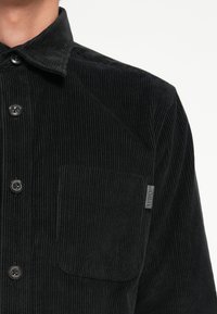 Schwarzes Cordhemd mit Knopfdesign, spitzem Kragen, Brusttasche und einem kleinen Stoffetikett an der Seite, das eine vertikale Rippstruktur zeigt.