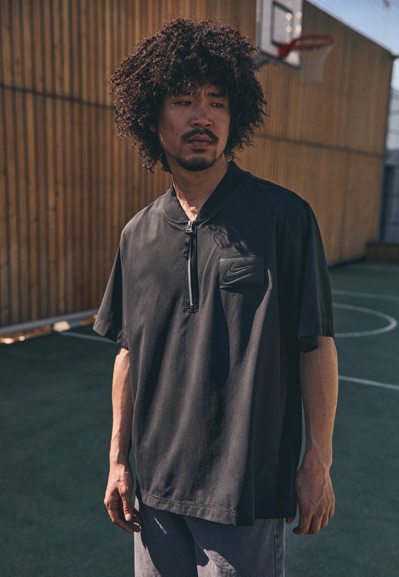 Sort Nike polo skjorte med halv lynlås og korte ærmer, med en forlommesyning prydet med Nike-logoen, lavet af letvægtsmateriale.