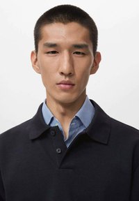 Polo negro con un collar abotonado superpuesto sobre una camisa a rayas azul y blanca. La tela parece suave, con un ajuste clásico y detalles mínimos.