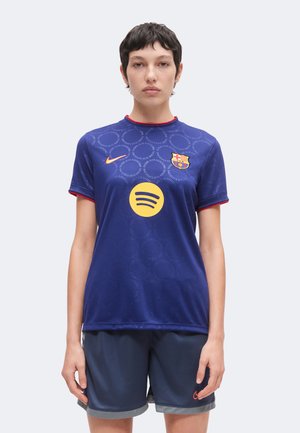 Persona de pie mirando al frente, vistiendo una camiseta azul del FC Barcelona con logotipos de patrocinadores y pantalones cortos deportivos azules, fondo neutro.