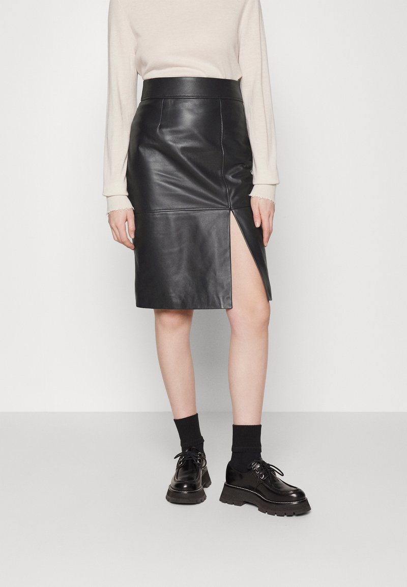 Zwarte leren kokerrok met een hoge split, gladde textuur, aansluitende tailleband, gestyled met zwarte schoenen en sokken. Eenvoudig, strak ontwerp.