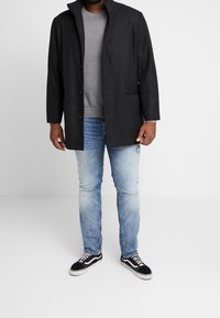 Cappotto nero con colletto alto, chiusura a singolo bottone, indossato sopra una camicia grigia, abbinato a jeans chiari strappati e sneakers nere.