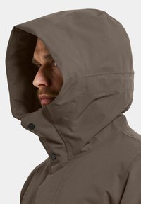 Ruskea vedenpitävä takki korkealla kauluksella ja suuressa hupussa, jossa on mustat painonapit ja sileä, mattapintainen rakenne.