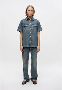 Denim outfit featuring a short-sleeve button-up shirt with two chest pockets and wide-leg jeans, all in a faded blue wash. Dark brown flats included.