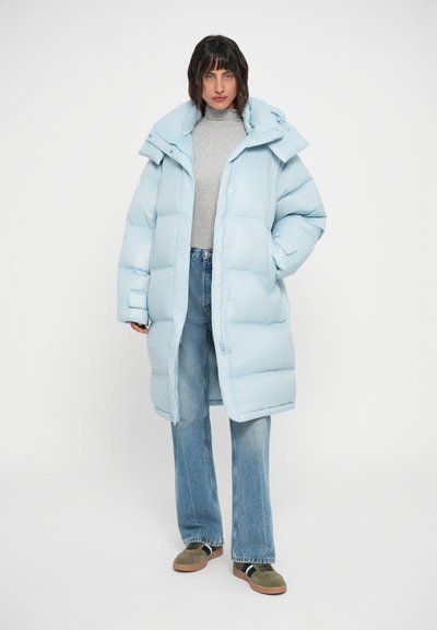 Gaiši zils pūkains mētelis ar augstu apkakli, pogām un sānu kabatām, kombinēts ar pelēku turtleneck un platām džinsu biksēm.