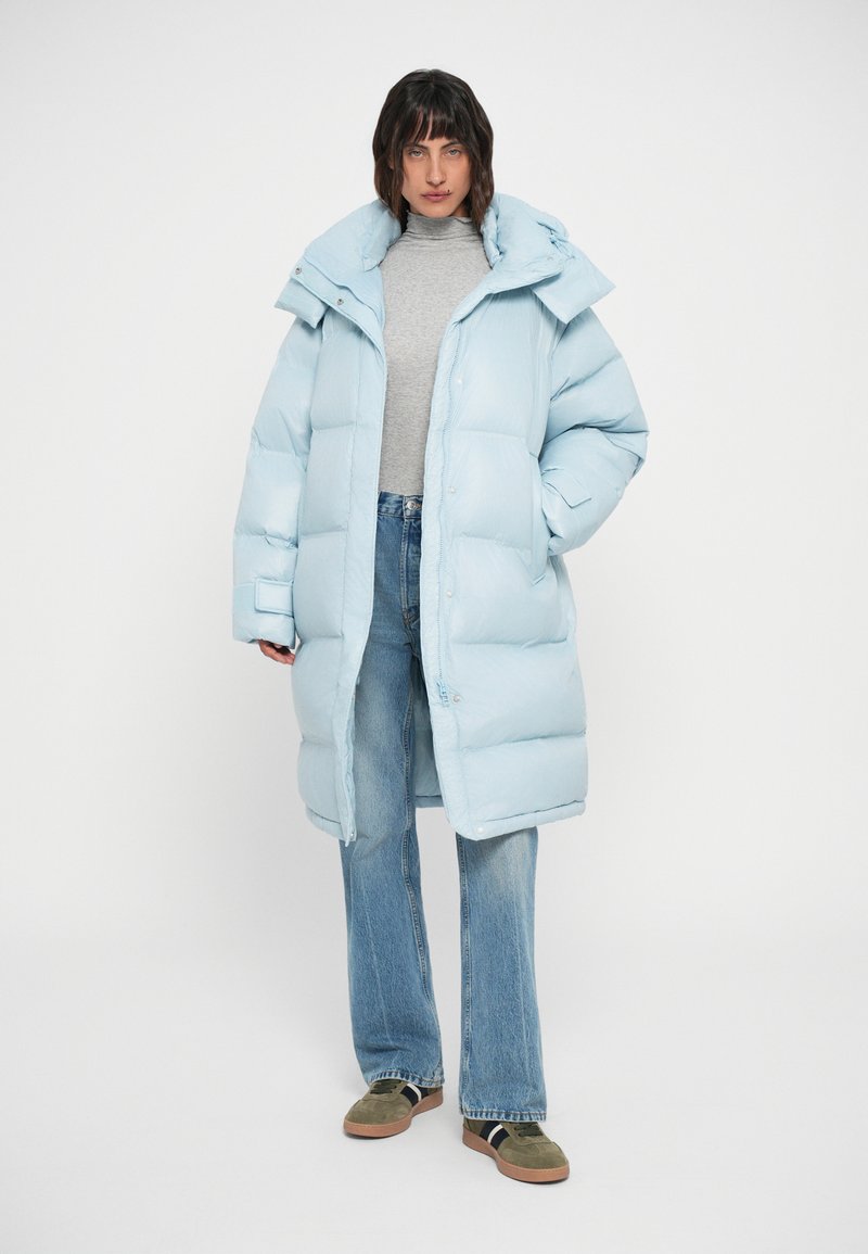 Manteau matelassé bleu clair avec un col haut, des boutons-pression et des poches latérales, associé à un pull à col roulé gris et un jean en denim évasé.