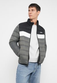 Polstret jakke i sort, grå og hvid med høj krave, lynlås lukning og horisontalt quiltet design. Båret over en lys grå sweatshirt.