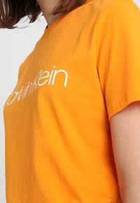 Camiseta de algodón naranja con un logo blanco de "Calvin Klein", manga corta y cuello redondo. Textura suave, ajuste clásico.