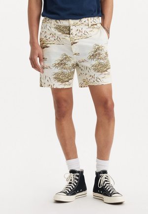 Homme portant un short beige à imprimé tropical, des chaussettes blanches et des baskets montantes noires, debout les mains dans les poches.