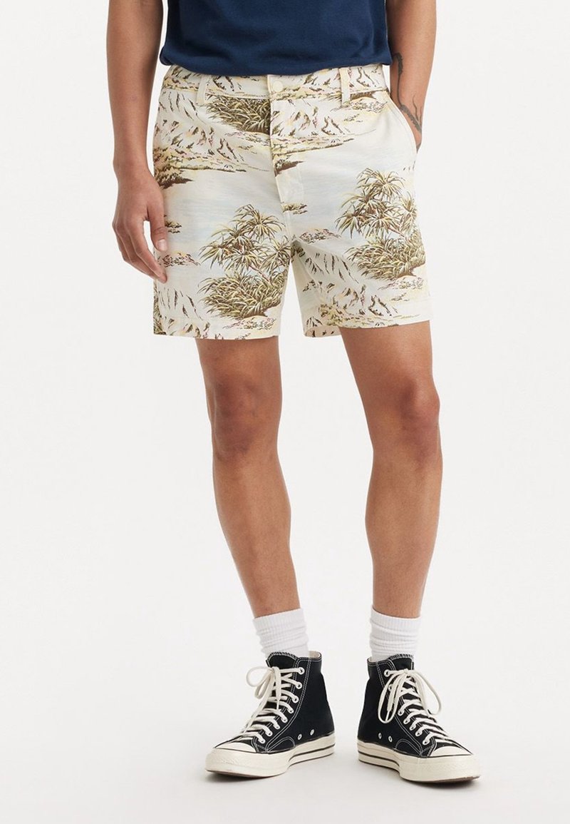 Homme portant un short beige à imprimé tropical, des chaussettes blanches et des baskets montantes noires, debout les mains dans les poches.