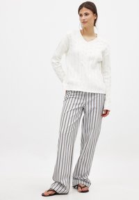 Weißer Kabelstrickpullover mit V-Ausschnitt, kombiniert mit gestreiften, weiten Beinkleidern in Beige, Grau und Weiß. Sandalen runden das Outfit ab.