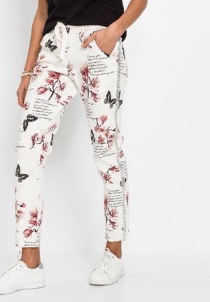 Donna che indossa pantaloni bianchi con stampa floreale, testo scritto a mano e farfalle nere, abbinati a sneakers bianche e una maglietta nera.