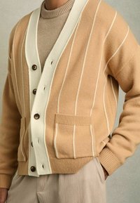 Cardigan beige à rayures avec un col châle blanc, des boutons marron et une poche avant, porté sur un pull léger marron. Tissu tricoté texturé.