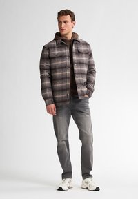 Grijze geruite flanellen shirtjack met twee borstzakken, gedragen over een bruine hoodie en gecombineerd met lichtgrijze jeans en witte sneakers.