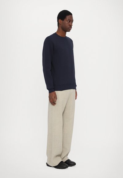 Sweatshirt marine à manches longues et poignets côtelés, associé à un pantalon large beige clair et des chaussures noires, porté par un modèle debout.