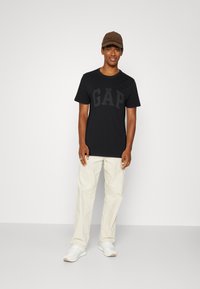 Schwarzes T-Shirt mit kurzen Ärmeln und dem fett gedruckten, strukturierten "GAP"-Logo. Kombiniert mit hellbeigen Hose und weißen Sneakers. Braune Kappe als Akzent.