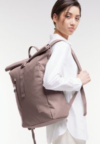 Zaino roll-top in tessuto mauve tenue, presenta spallacci regolabili, una tasca con zip laterale e un accento logo minimale sulla parte anteriore.
