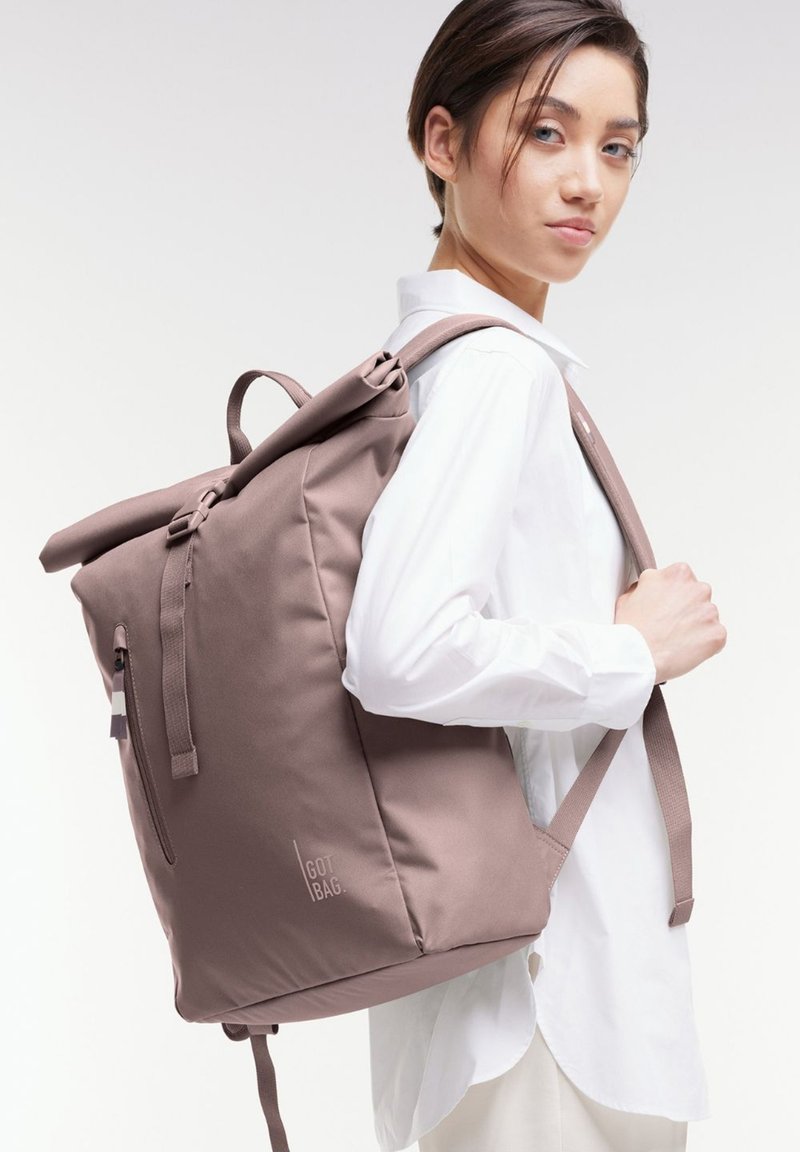 Zaino roll-top in tessuto mauve tenue, presenta spallacci regolabili, una tasca con zip laterale e un accento logo minimale sulla parte anteriore.