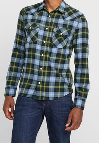 Rutigt skjorta med en blandning av blå, grön och gul rutnät, långa ärmar, spetsig krage och två bröstfickor med tryckknappar.