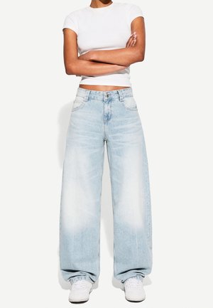 Persona che indossa una t-shirt bianca corta, jeans ampi azzurri e sneaker bianche con le braccia incrociate.