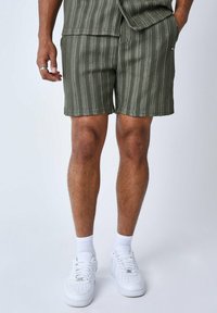 Pantaloni corti verde oliva con strisce verticali bianche, realizzati in tessuto testurizzato. Indossati con scarpe da ginnastica bianche e calze alla caviglia.