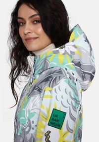 Färgglad mönstrad huva jacka i ljusblått, med blommönster i grått och gult, samt en grön Urban Wear-logotyp på ärmen.
