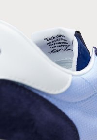 Buty sportowe z gładką białą nakładką na piętę, jasnoniebieską siateczkową cholewką oraz ciemnoniebieskimi zamszowymi akcentami. Wewnątrz znajduje się etykieta z motywującym cytatem.