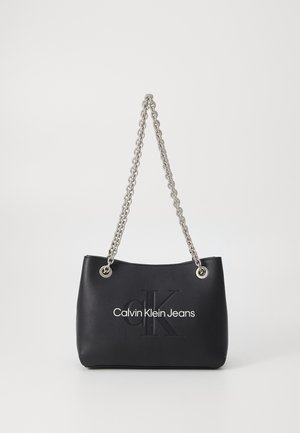 Sort Calvin Klein håndtaske med sølvkæderem og præget "CK" logo foran mod en ensartet lys baggrund.